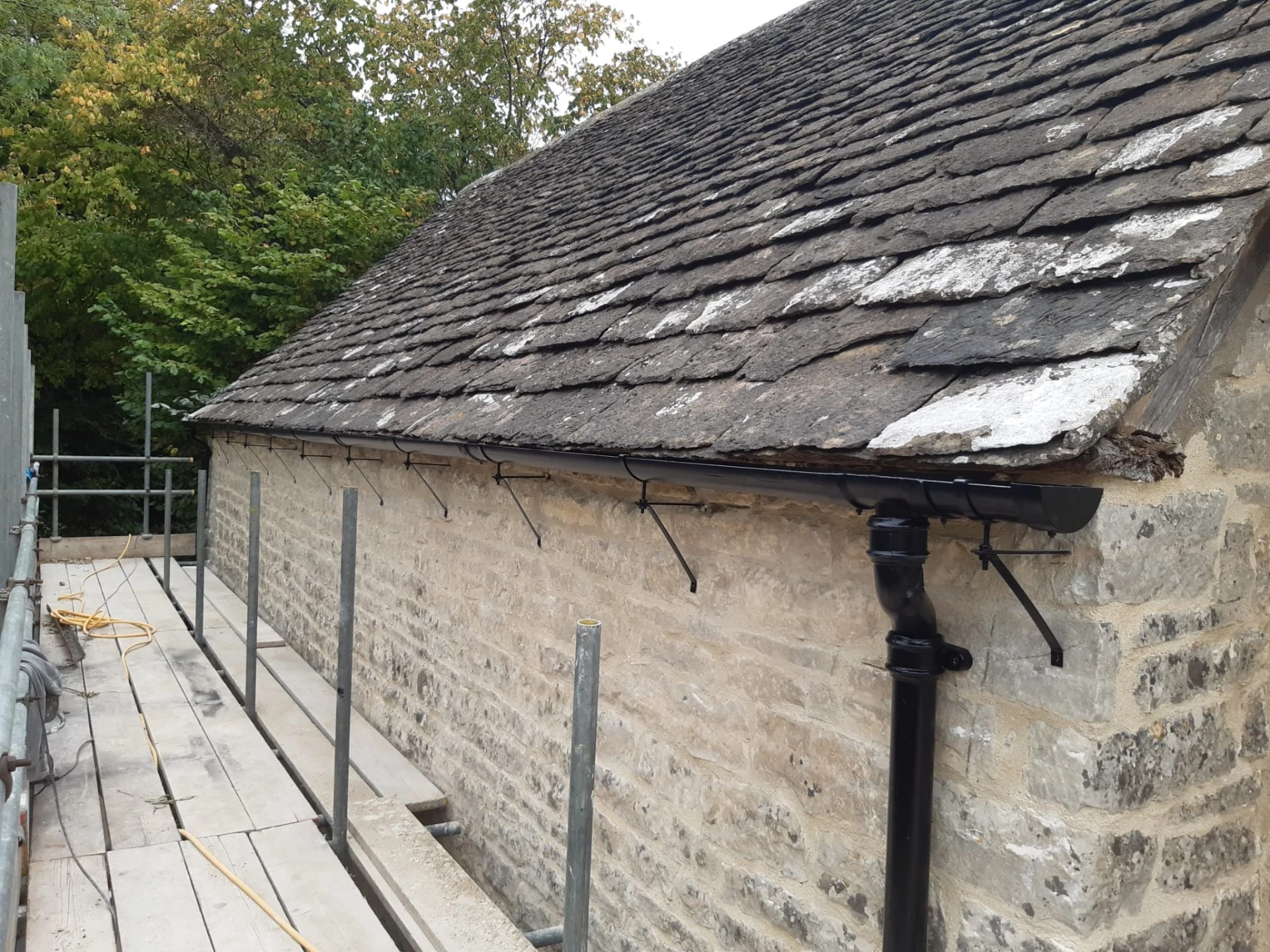 Black guttering with supporting brackets and downpipe outlet beneath a traditional stone-tiled roof