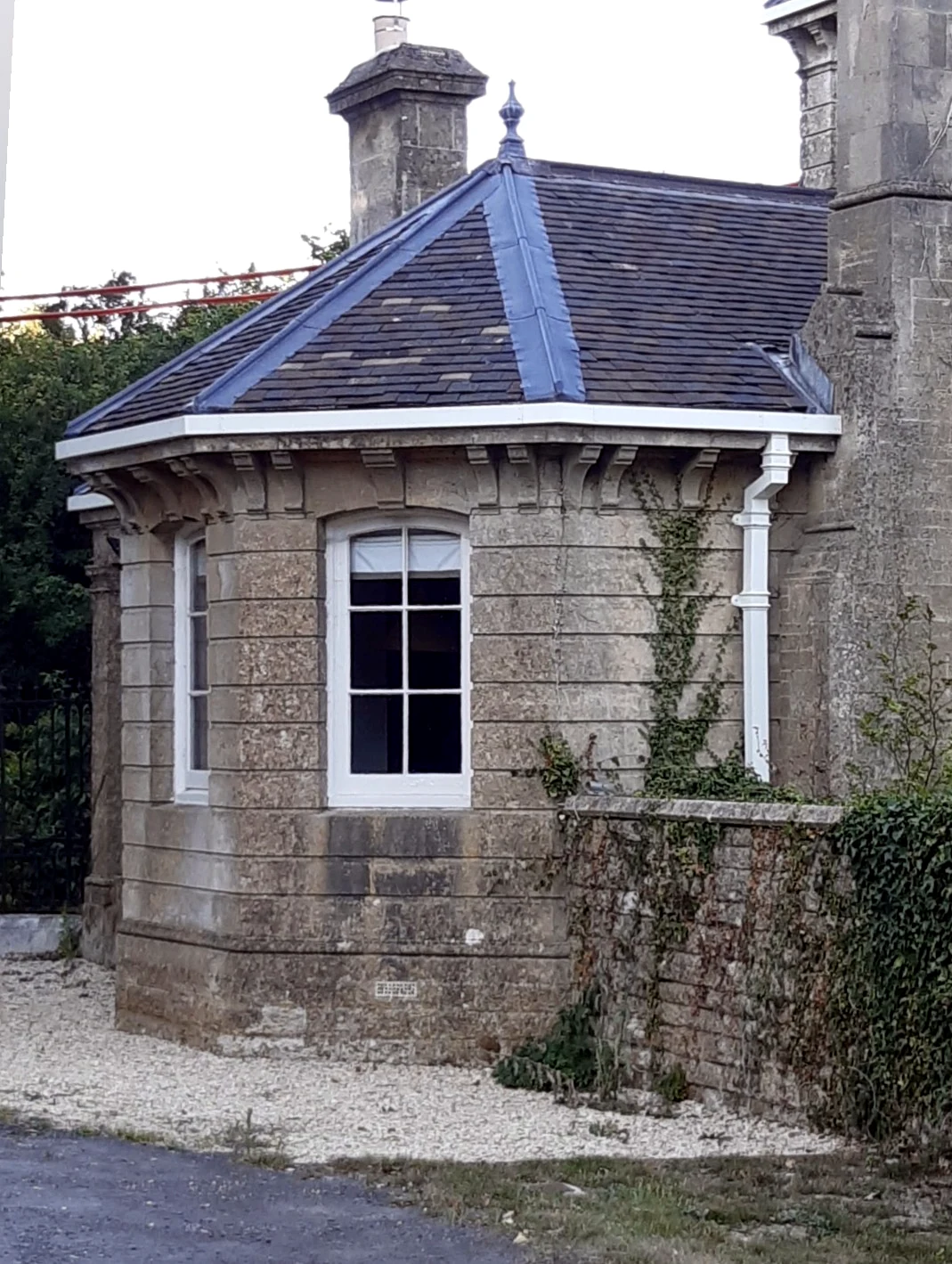 White guttering and downpipe beneath a bracketed stone cornice on a period stone lodge with a faceted bay window and slate hipped roof
